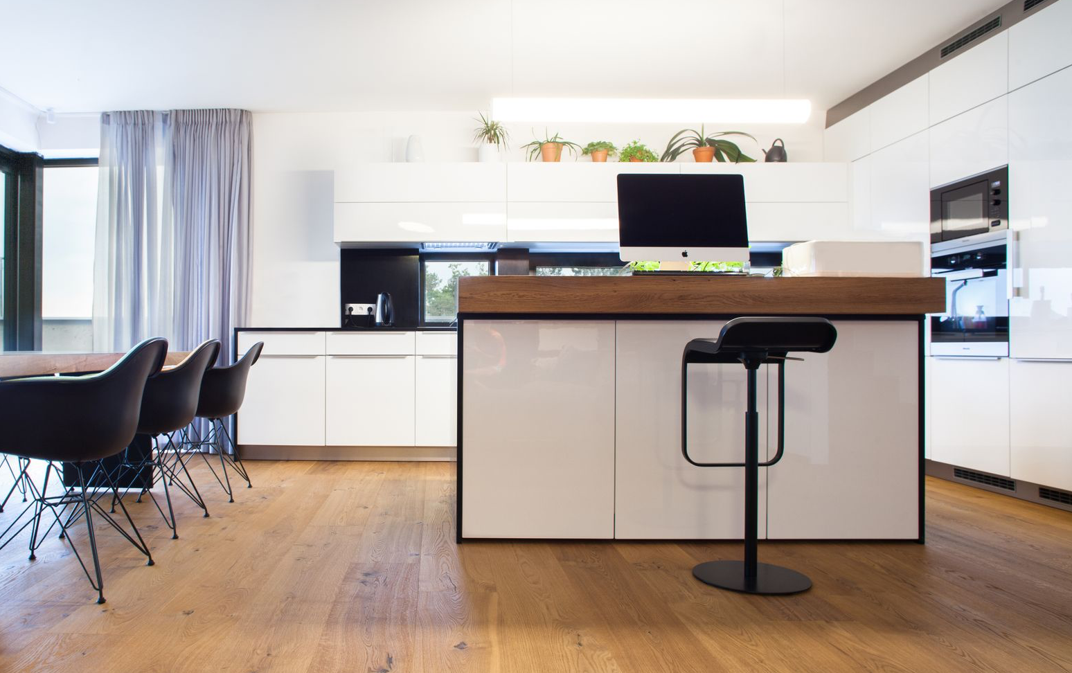Moderní bílá kuchyně s dřevěným ostrůvkem a dubovou podlahou v teplém odstínu, doplněná černými designovými židlemi a prosklenou stěnou s výhledem.
